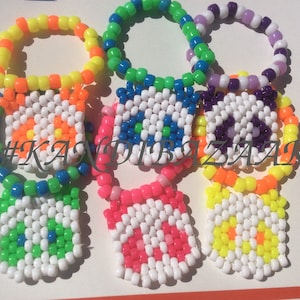 Puede incluir: Cinco pulseras de cuentas de colores con dijes cuadrados. Los dijes son blancos con un dise&ntilde;o colorido en el centro. Las pulseras est&aacute;n hechas de cuentas de color amarillo ne&oacute;n, naranja, verde, azul y morado. Los dijes tienen dise&ntilde;os de una flor, un ojo, un signo de paz, una flor y una flor.