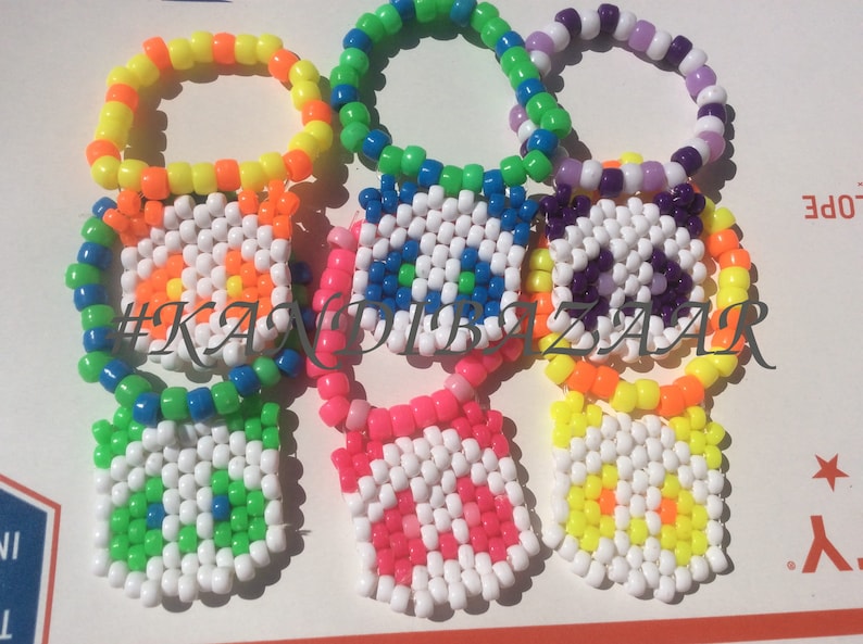Puede incluir: Cinco pulseras de cuentas de colores con dijes cuadrados. Los dijes son blancos con un dise&ntilde;o colorido en el centro. Las pulseras est&aacute;n hechas de cuentas de color amarillo ne&oacute;n, naranja, verde, azul y morado. Los dijes tienen dise&ntilde;os de una flor, un ojo, un signo de paz, una flor y una flor.
