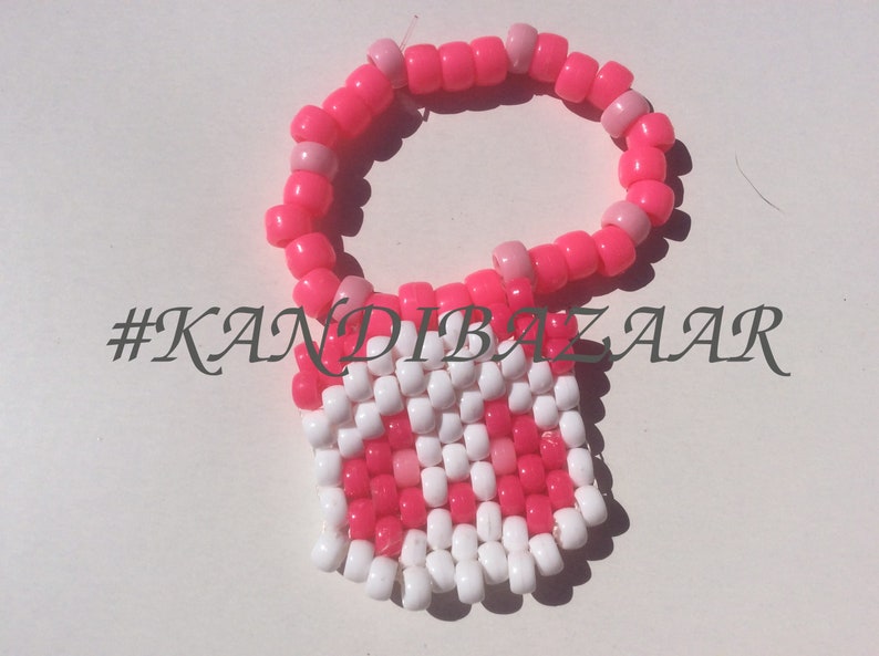 Puede incluir: Pulsera kandi rosa y blanca con un encanto cuadrado. El encanto est&aacute; hecho de cuentas rosas y blancas y tiene un patr&oacute;n de cuadros. La pulsera tiene el hashtag #KANDIBAZAAR.
