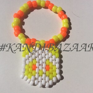 Puede incluir: Un brazalete kandi amarillo y naranja ne&oacute;n con un dije de cuentas blancas, amarillas y naranjas. El dije tiene un dise&ntilde;o simple con algunas cuentas naranjas en el centro. El brazalete tiene el hashtag #KANDIBAZAAR.