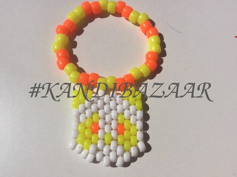 Puede incluir: Un brazalete kandi amarillo y naranja ne&oacute;n con un dije de cuentas blancas, amarillas y naranjas. El dije tiene un dise&ntilde;o simple con algunas cuentas naranjas en el centro. El brazalete tiene el hashtag #KANDIBAZAAR.