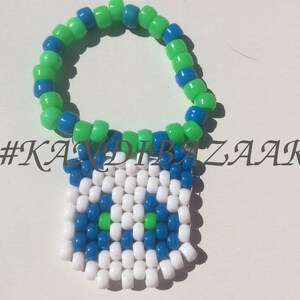 Puede incluir: Un brazalete de cuentas verde y azul con un dise&ntilde;o pixelado blanco, azul y verde. El dise&ntilde;o es una cara con dos ojos y una boca. El hashtag #KANDIBZAR es visible en la imagen.
