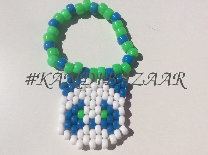 Puede incluir: Un brazalete de cuentas verde y azul con un dise&ntilde;o pixelado blanco, azul y verde. El dise&ntilde;o es una cara con dos ojos y una boca. El hashtag #KANDIBZAR es visible en la imagen.