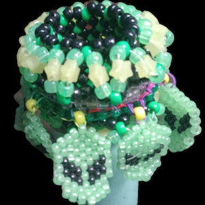 May include: A multi-layered beaded bracelet with green, black, and yellow beads. The bracelet is made of small, round beads and features a star-shaped design. The bracelet is decorated with several small, green charms with black eyes.