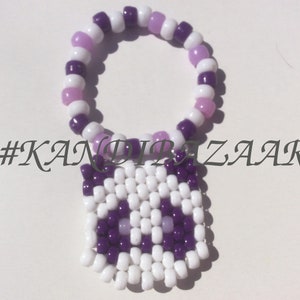 Puede incluir: Un brazalete de cuentas blancas y moradas con un dije de b&uacute;ho blanco y morado. El brazalete est&aacute; hecho con cuentas peque&ntilde;as y redondas, y el dije est&aacute; hecho con cuentas cuadradas. El dije tiene una cara blanca con ojos morados y un pico morado. El brazalete est&aacute; etiquetado con el hashtag #KANDIBAZAAR.