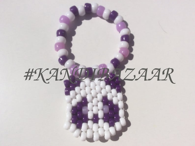 Puede incluir: Un brazalete de cuentas blancas y moradas con un dije de b&uacute;ho blanco y morado. El brazalete est&aacute; hecho con cuentas peque&ntilde;as y redondas, y el dije est&aacute; hecho con cuentas cuadradas. El dije tiene una cara blanca con ojos morados y un pico morado. El brazalete est&aacute; etiquetado con el hashtag #KANDIBAZAAR.