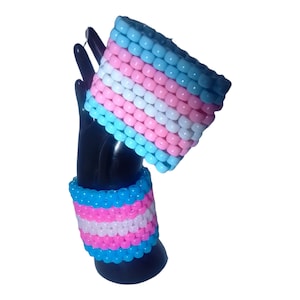 May include: Two beaded bracelets in the colors of the transgender pride flag. The bracelets are made of small, colorful beads and are stacked on top of each other.