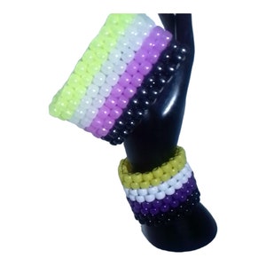 May include: Two beaded bracelets in the colors of the non-binary pride flag. The top bracelet is yellow, white, pink, and black. The bottom bracelet is yellow, white, purple, and black.