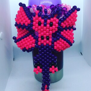 Op de afbeelding: Een roze en paarse kralen drakenbedel met een lange staart. De bedel is gemaakt van kleine, kleurrijke kralen en is een populair accessoire voor sleutelhangers, tassen en sieraden.