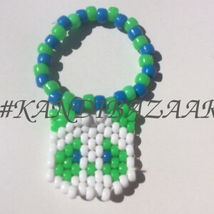 Puede incluir: Un brazalete kandi verde, azul y blanco con un amuleto cuadrado. El amuleto est&aacute; hecho de cuentas blancas, verdes y azules y tiene un dise&ntilde;o pixelado. El brazalete est&aacute; etiquetado con el hashtag #KANDIBAZAAR.