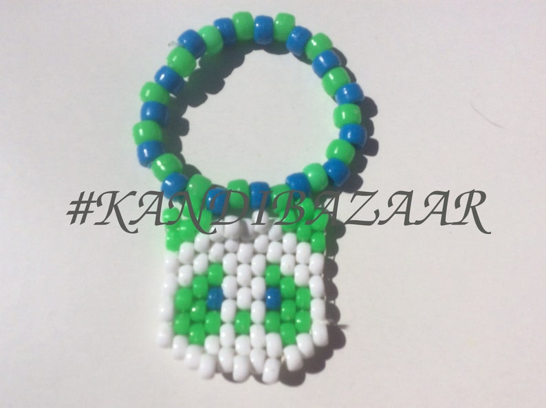 Puede incluir: Un brazalete kandi verde, azul y blanco con un amuleto cuadrado. El amuleto est&aacute; hecho de cuentas blancas, verdes y azules y tiene un dise&ntilde;o pixelado. El brazalete est&aacute; etiquetado con el hashtag #KANDIBAZAAR.