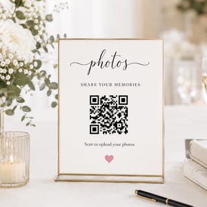 May include: A rectangular sign with a thin gold frame featuring the word photos in black script, the text SHARE YOUR MEMORIES, a black and white QR code, the text Scan to upload your photos, and a small pink heart at the bottom center.