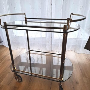 Peut inclure: Chariot de service vintage avec deux étagères ovales en verre et des accents en métal couleur laiton. Le chariot a quatre roues et des poignées incurvées. Le cadre est de couleur marron foncé.