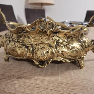 May include: A gold-toned metal planter with intricate floral designs and scrollwork. The planter has four feet and two handles.
