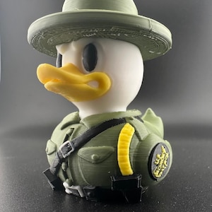 May include: A 3D-printed duck figurine wearing a green ranger hat and uniform. The duck has a white head, black eyes, and a yellow beak. The uniform includes a black strap and a yellow patch.