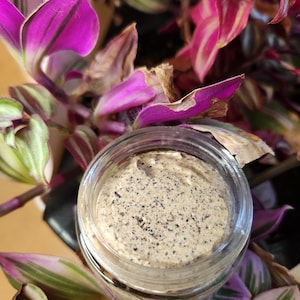 May include: A clear glass jar filled with a beige, speckled cosmetic product. The jar sits amidst vibrant purple and green plant leaves, creating a natural, botanical aesthetic. The product appears to be a scrub or mask.
