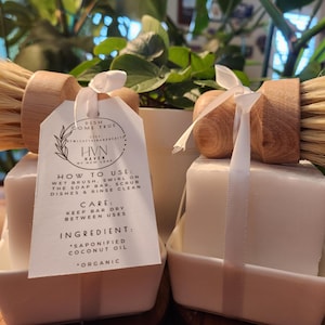 May include: Two square white soap bars in white ceramic dishes, each topped with a round wooden dish brush with light-colored bristles. A white tag tied with a ribbon reads: DISH COME TRUE, HVN HAVEN BY NEW LEAF, HOW TO USE, CARE, INGREDIENT.