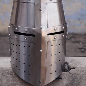 Peut inclure: Casque de chevalier en acier poli avec une construction rivet&eacute;e. Le casque pr&eacute;sente un dessus plat, une croix sur le devant et des fentes horizontales pour les yeux. De nombreux petits trous sont pr&eacute;sents sur la partie inf&eacute;rieure.