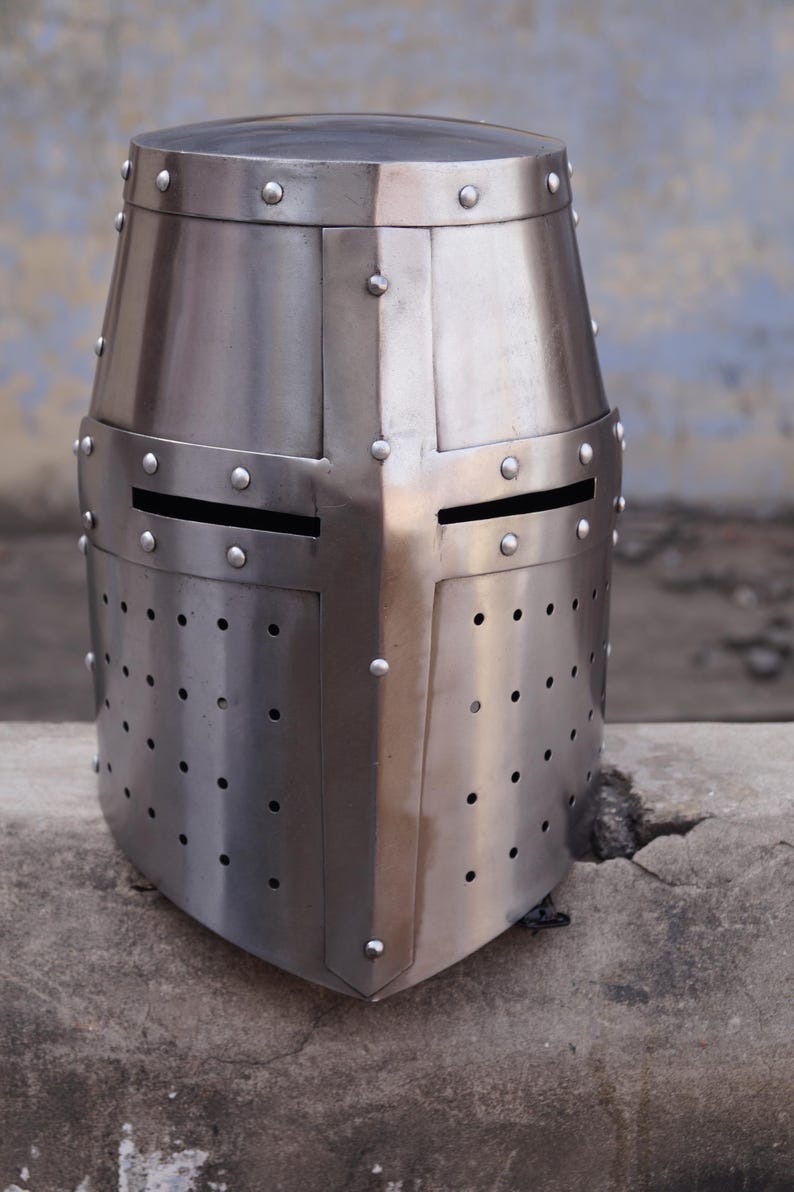 Peut inclure: Casque de chevalier en acier poli avec une construction rivet&eacute;e. Le casque pr&eacute;sente un dessus plat, une croix sur le devant et des fentes horizontales pour les yeux. De nombreux petits trous sont pr&eacute;sents sur la partie inf&eacute;rieure.