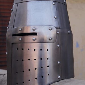 Peut inclure: Casque de style m&eacute;di&eacute;val argent&eacute;, avec un dessus plat et une plaque frontale rectangulaire. La plaque frontale comporte de nombreux petits trous espac&eacute;s et une fente horizontale pour les yeux. Des rivets fixent les panneaux du casque.