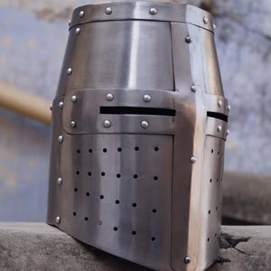 Peut inclure: Casque de style m&eacute;di&eacute;val de couleur argent&eacute;e, avec un dessus plat et une construction rivet&eacute;e. Le casque pr&eacute;sente une plaque frontale rectangulaire avec des fentes horizontales pour les yeux et de nombreux trous de ventilation. Le design rappelle une armure historique.
