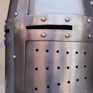 Peut inclure: Gros plan d'un casque m&eacute;di&eacute;val argent&eacute; et poli. Le casque pr&eacute;sente une fente oculaire rectangulaire et de nombreux rivets. Des rang&eacute;es de petits trous espac&eacute;s sont visibles sur la partie inf&eacute;rieure, sugg&eacute;rant une ventilation. La surface m&eacute;tallique refl&egrave;te la lumi&egrave;re.