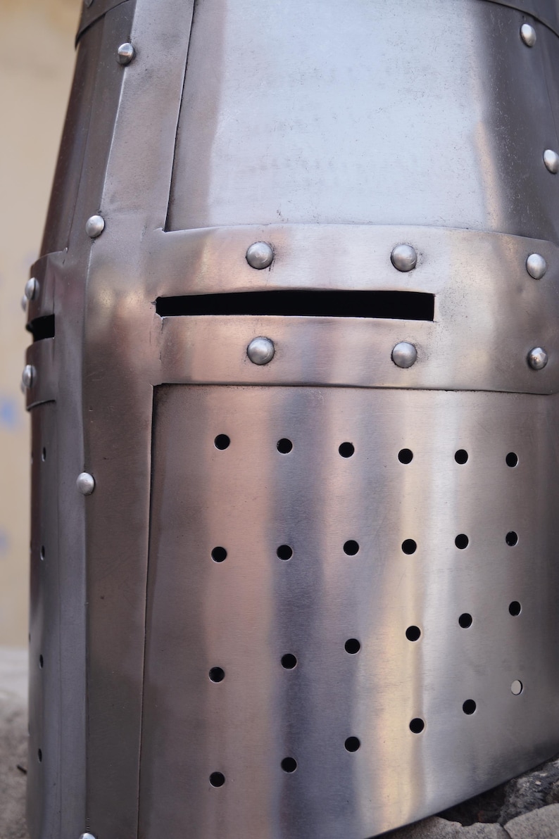 Peut inclure: Gros plan d'un casque m&eacute;di&eacute;val argent&eacute; et poli. Le casque pr&eacute;sente une fente oculaire rectangulaire et de nombreux rivets. Des rang&eacute;es de petits trous espac&eacute;s sont visibles sur la partie inf&eacute;rieure, sugg&eacute;rant une ventilation. La surface m&eacute;tallique refl&egrave;te la lumi&egrave;re.
