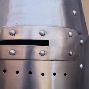Peut inclure: Gros plan sur un casque de chevalier m&eacute;di&eacute;val en acier poli. Le casque comporte une fente oculaire rectangulaire et est rivet&eacute;. Le m&eacute;tal a une surface r&eacute;fl&eacute;chissante, et le design est simple et fonctionnel.