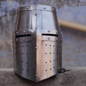 Peut inclure: Casque de chevalier argent&eacute;, en m&eacute;tal rivet&eacute;. Il pr&eacute;sente un design en croix, des fentes pour les yeux et de petits trous de ventilation. La surface m&eacute;tallique refl&egrave;te la lumi&egrave;re, mettant en valeur son design historique.