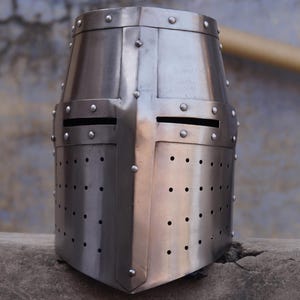 Peut inclure: Casque de crois&eacute; m&eacute;di&eacute;val argent&eacute; et poli. Le casque pr&eacute;sente un dessus plat, un motif en forme de croix et de petits rivets. Il comporte des fentes pour les yeux et de nombreux trous de ventilation.