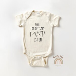 May include: Cream-colored baby onesie with the text "Shhh... Daddy Says Math Is Fun" in black. The onesie has short sleeves and a snap closure. A small rainbow graphic and the words "Gentle Hug Babywear" are at the bottom.