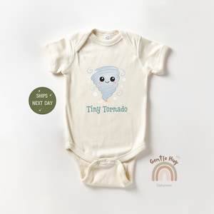 May include: A cream-colored baby onesie with a cartoon tornado design. The tornado is blue and gray with a smiling face. The text "Tiny Tornado" is printed below the tornado.