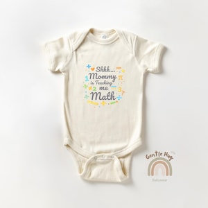 May include: Cream-colored baby bodysuit with short sleeves. The front features colorful text that reads "Shhh... Mommy is Teaching me Math" with math symbols and a rainbow graphic. The bodysuit is made of soft material.