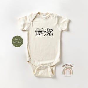 May include: A cream-colored baby onesie with short sleeves and a black graphic that reads "Relax, My Daddy's a Welder". The graphic includes a cartoon image of a welder.