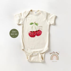 May include: Cream-colored baby onesie featuring a cheerful cherry graphic with the text "sweet like a cherry." The illustration shows two smiling cherries with green leaves. The onesie has short sleeves and a snap closure.