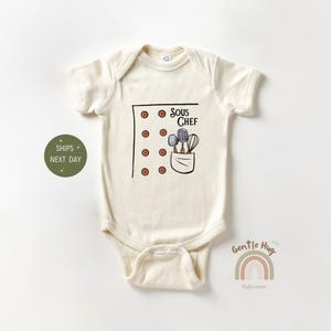 May include: A white baby onesie with a brown pocket and the text "Sous Chef" in black. The pocket has a spatula, a spoon, and a whisk in it. There are 10 brown buttons on the onesie.