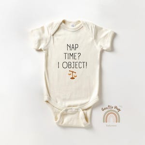 May include: Cream-colored baby bodysuit with the text "NAP TIME? I OBJECT!" in black, and a small scale illustration. The bodysuit has short sleeves and a snap closure. A rainbow logo and the words "Gentle Hug Babywear" are at the bottom.