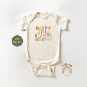 May include: A cream-colored baby onesie with the text "Mama's Little Reading Buddy" in colorful, rounded letters. A stack of books is pictured next to the text. The onesie is from the brand "Gentle Hug Babywear."