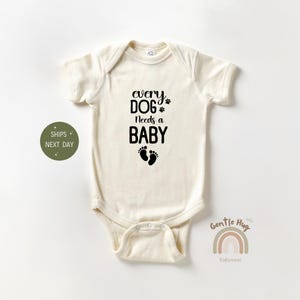 May include: A white baby onesie with black text that reads "every DOG needs a BABY" with two black paw prints above the word "DOG" and two black footprints below the word "BABY".