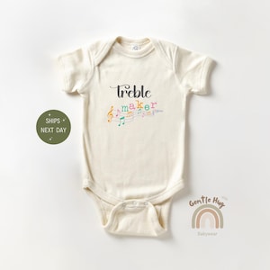 May include: A cream-colored baby onesie with short sleeves and a snap closure. The onesie has a black graphic that says "Treble Maker" with colorful musical notes.