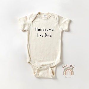May include: Cream-colored baby onesie with the text "Handsome like Dad" in black. The onesie has short sleeves and a snap closure. A small rainbow graphic and the words "Gentle Hug Babywear" are at the bottom.