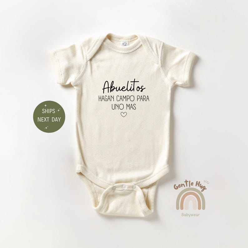 K&ouml;nnte beinhalten: Ein wei&szlig;er Baby-Strampler mit schwarzem Text, der "Abuelitos Hagan Campo Para Uno Mas" mit einem kleinen Herzsymbol lautet.