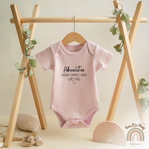 K&ouml;nnte beinhalten: Rosafarbener Baby-Strampler mit kurzen &Auml;rmeln und dem Text "Abuelitos hagan campo para uno mas" in Schwarz.