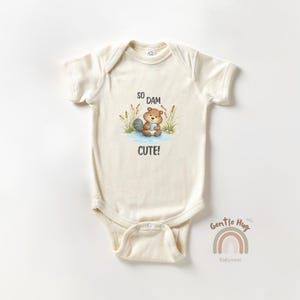May include: Cream-colored baby onesie with a beaver illustration and the text "So Dam Cute!". The beaver is in a pond with cattails. The onesie has short sleeves and a snap closure.