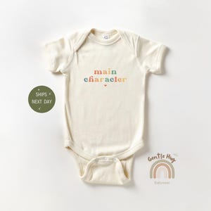 May include: A cream-colored baby onesie with the text "main character" in rainbow colors. The onesie is from Gentle Hug Babywear.