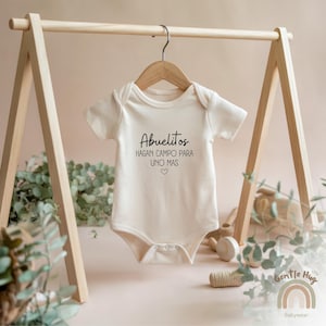 K&ouml;nnte beinhalten: Ein wei&szlig;er Baby-Strampler mit dem Text "Abuelitos hagan campo para uno mas" in schwarzer Schrift. Der Strampler h&auml;ngt an einem Holzb&uuml;gel.