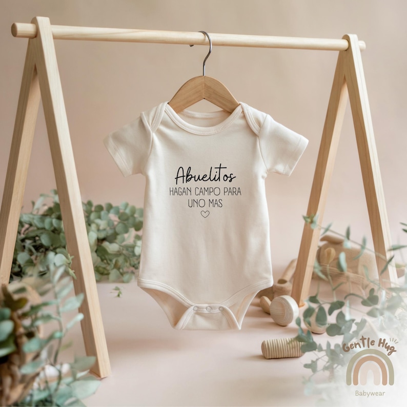 K&ouml;nnte beinhalten: Ein wei&szlig;er Baby-Strampler mit dem Text "Abuelitos hagan campo para uno mas" in schwarzer Schrift. Der Strampler h&auml;ngt an einem Holzb&uuml;gel.