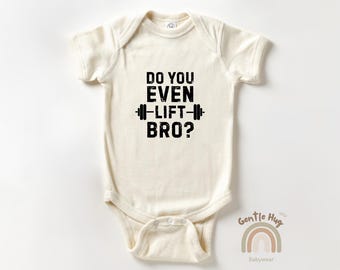 May include: Cream-colored baby onesie with the text "DO YOU EVEN LIFT BRO?" in black, along with a barbell graphic. The onesie has short sleeves and a snap closure. The Gentle Hug Babywear logo is in the bottom right corner.