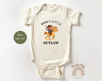 May include: Cream-colored baby bodysuit with a cowboy graphic. The illustration depicts a cowboy riding a horse, with the text "Mama's Little Outlaw" above and "Outlaw" below the graphic. The bodysuit is made of soft material.