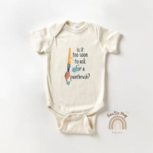 May include: Cream-colored baby bodysuit with the text "is it too soon to ask for a paintbrush?" and a watercolor paintbrush illustration. The design includes blue paint splatters. The Gentle Hug Babywear logo is at the bottom.